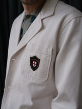 A person wears the BGbaio Twill Blazer in Chalk by Blue de Gênes, featuring a chest pocket crest with a shield and red cross. The blazer’s regular fit partly conceals their face, which is out of frame.