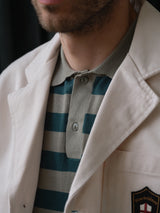 A close-up of a man in the Blue de Gênes BGbaio Twill Blazer in Chalk over a gray and green striped polo, with part of his face, beard, and the blazer's embroidered patch visible.