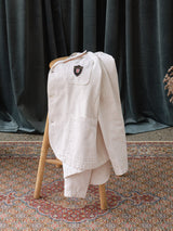 The BGbaio Twill Blazer - Chalk by Blue de Gênes, a cream cotton jacket with a chest patch, rests on a wooden chair atop an ornate rug with dark curtains behind.
