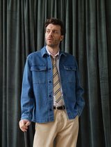 A man in a Blue de Gênes BGpontus Denim Overshirt - Used, white shirt, striped tie, and beige pants stands before dark green curtains, cane in left hand, looking slightly to the side.