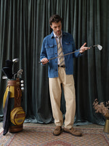 A man stands indoors on a patterned rug, wearing a denim jacket, beige pants, and a Blue de Gênes BGponsy Tie in pale khaki. He holds a golf club; beside him are a golf bag with clubs and a yellow bag. A dark green curtain is behind him.