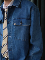 A person wears the Blue de Gênes BGpontus Denim Overshirt - Used as an overshirt, layered over a white shirt and a beige tie with navy stripes, upper torso visible against a dark background.