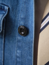 Close-up of the BGpontus Denim Overshirt - Used by Blue de Gênes in blue cotton blend with a black button engraved "rue de cette." A beige and black striped necktie appears partially in the background.