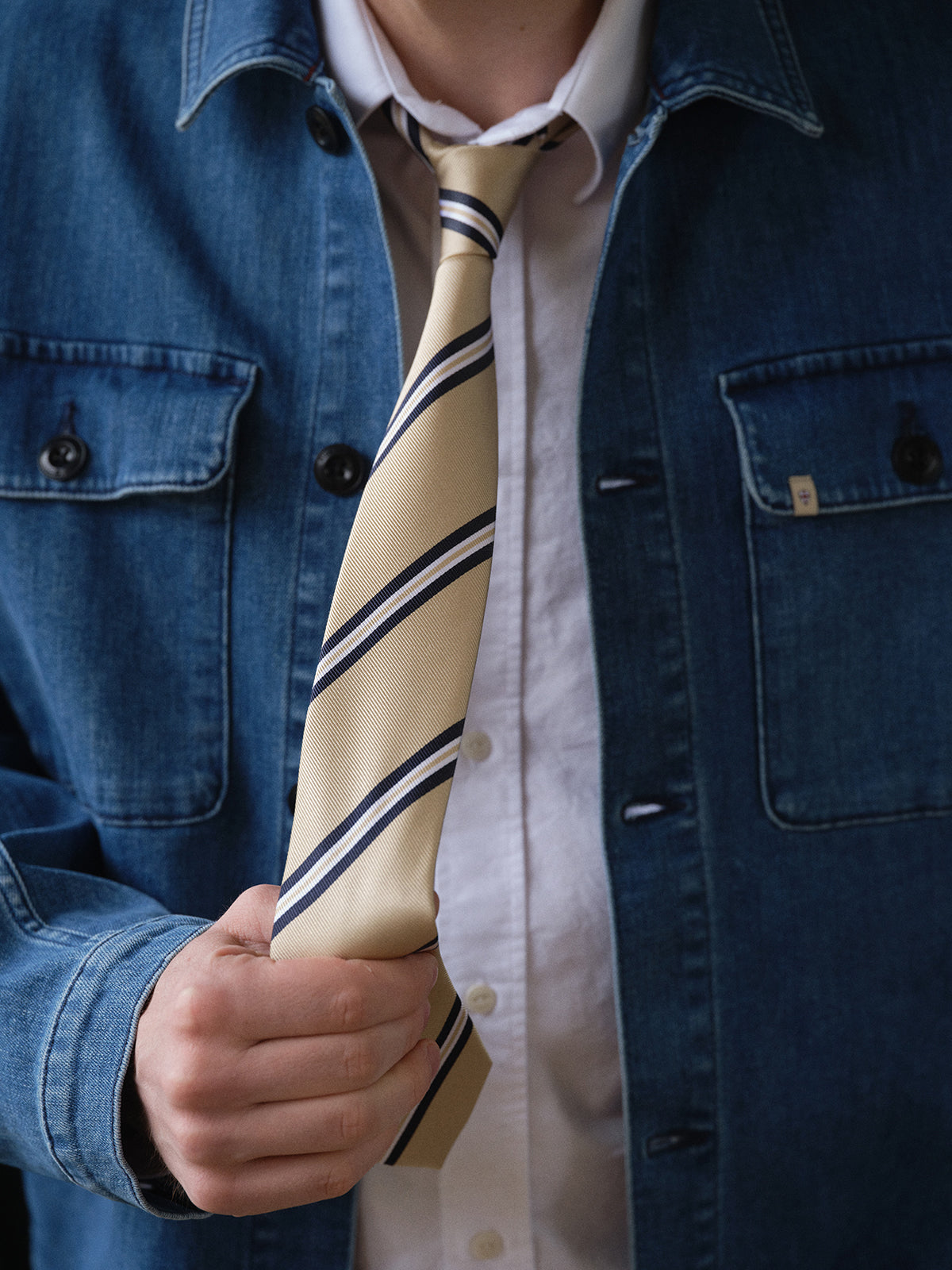 A person in a white shirt and blue denim jacket holds the end of a Blue de Gênes BGponsy Tie - Pale Khaki, loosely tied around their neck.