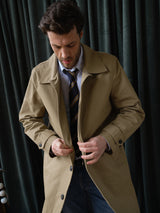 A man with dark hair stands before dark green curtains, looking down as he buttons the Blue de Gênes BGjacques Coat in Pale Khaki, worn over a blue striped shirt and tie.
