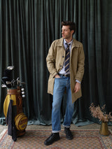 A man in a tan trench coat, striped tie, blue shirt, and Blue de Gênes BGvinci Hera Used Jeans stands on a patterned rug before dark green curtains. Nearby are a golf bag with clubs and a small vase of dried flowers.