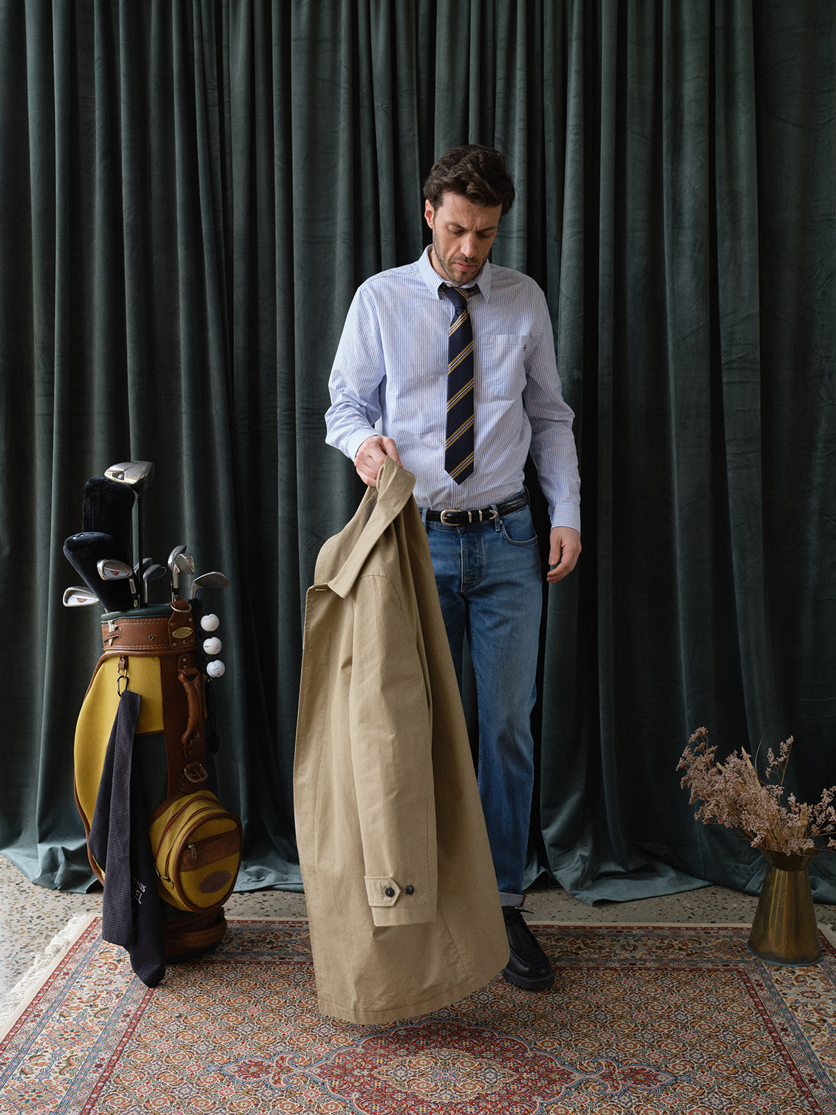 A man in classic workwear—a dress shirt, striped tie, and jeans—holds the BGjacques Coat in pale khaki by Blue de Gênes, standing on a patterned rug near a golf bag with clubs and dried flowers, set against dark green curtains.