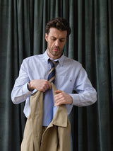 A man wearing the BGsander Oxford Shirt - Blue Stripe by Blue de Gênes stands before dark green curtains, looking down while holding a beige coat.