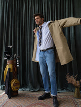 A man in Blue de Gênes BGvinci Hera Used Jeans, a striped tie, and a beige trench coat puts on his jacket next to a golf bag with clubs, standing on a patterned rug before dark green curtains.