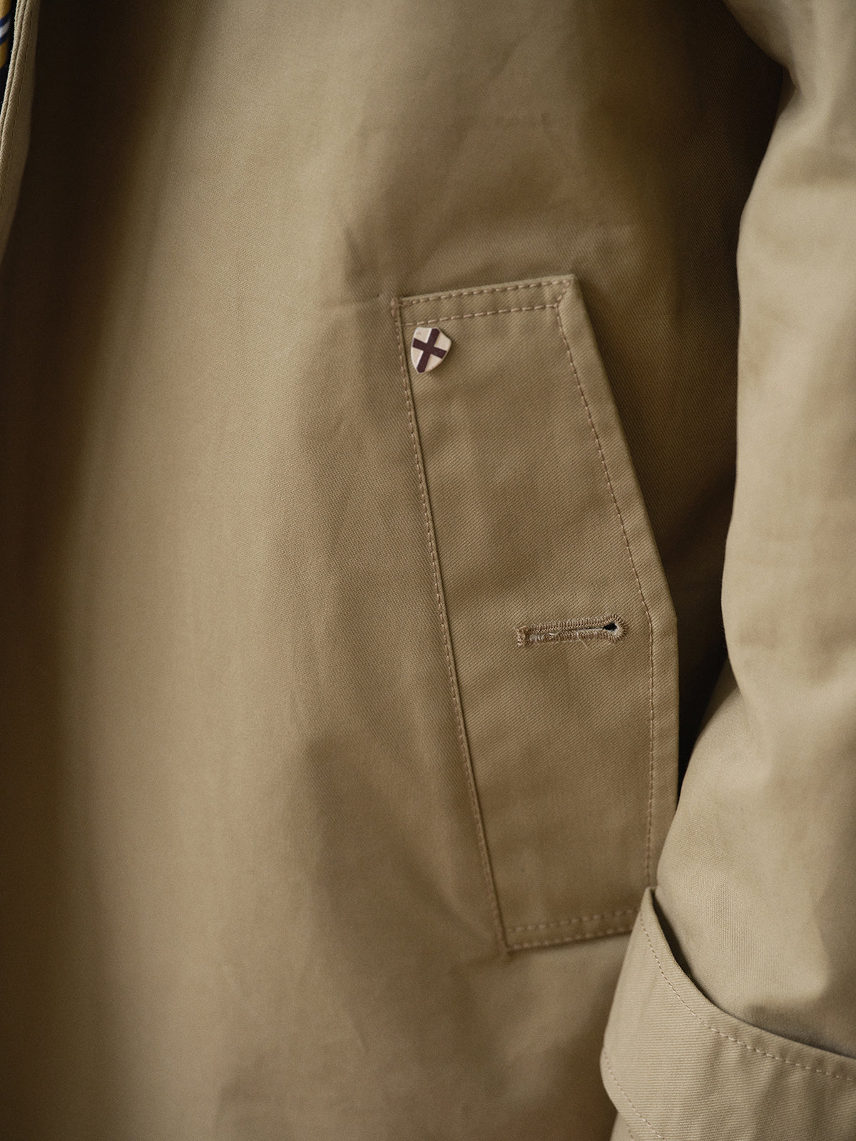 Close-up of the BGjacques Coat in Pale Khaki by Blue de Gênes, showing a heavy cotton pocket with a small red cross pin on a white background near the top. The sleeve and part of the coat’s body are also visible.