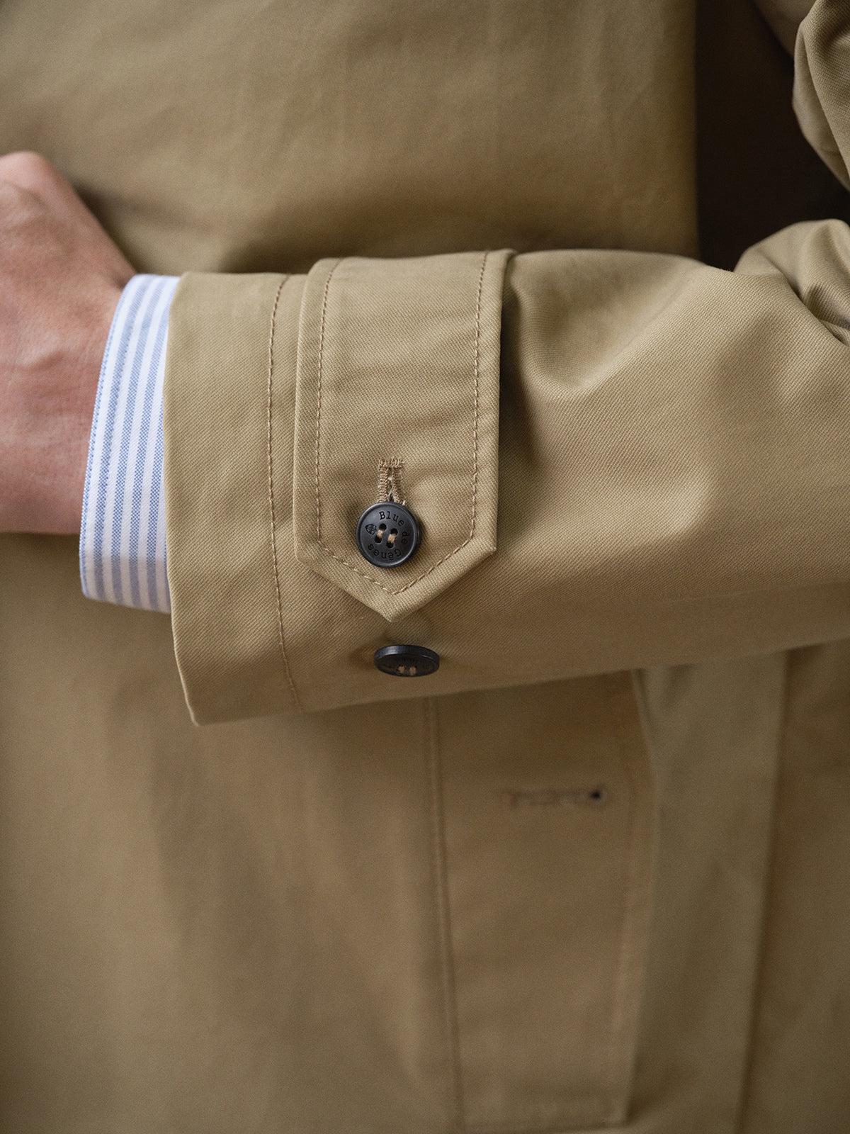 A close-up of a person's arm wearing the BGjacques Coat in Pale Khaki by Blue de Gênes, featuring black cuff buttons and a blue and white striped shirt sleeve underneath—evoking timeless workwear style.
