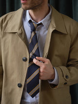 A man in a tan trench coat over a Blue de Gênes blue and white striped shirt adjusts his BGponsy Tie - Dark Navy by Blue de Gênes. His face is partly visible, showing a neatly trimmed beard and mustache.