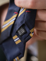 A close-up of a hand holding the BGponsy Tie - Dark Navy by Blue de Gênes, featuring navy stripes with yellow and white accents. The label displays "100% silk" in English, French, German, and Italian.