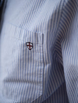 Close-up of the BGsander Oxford Shirt - Blue Stripe by Blue de Gênes, showing its light blue and white striped pocket with a small shield patch. Made from 100% cotton and finished with a classic button-down collar.