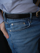 A person wearing Blue de Gênes BGvinci Hera Used Jeans in a used wash, styled with a black belt and a blue-and-white striped shirt. Their left hand is resting in the pocket of the comfort stretch denim jeans.