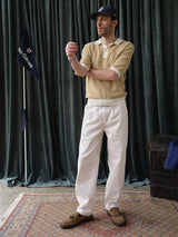 A man in a striped yellow polo, Blue de Gênes BGpalco Twill Pants in Chalk, brown shoes, and a navy cap stands on a patterned rug near dark green curtains, beside a flag and trunk.
