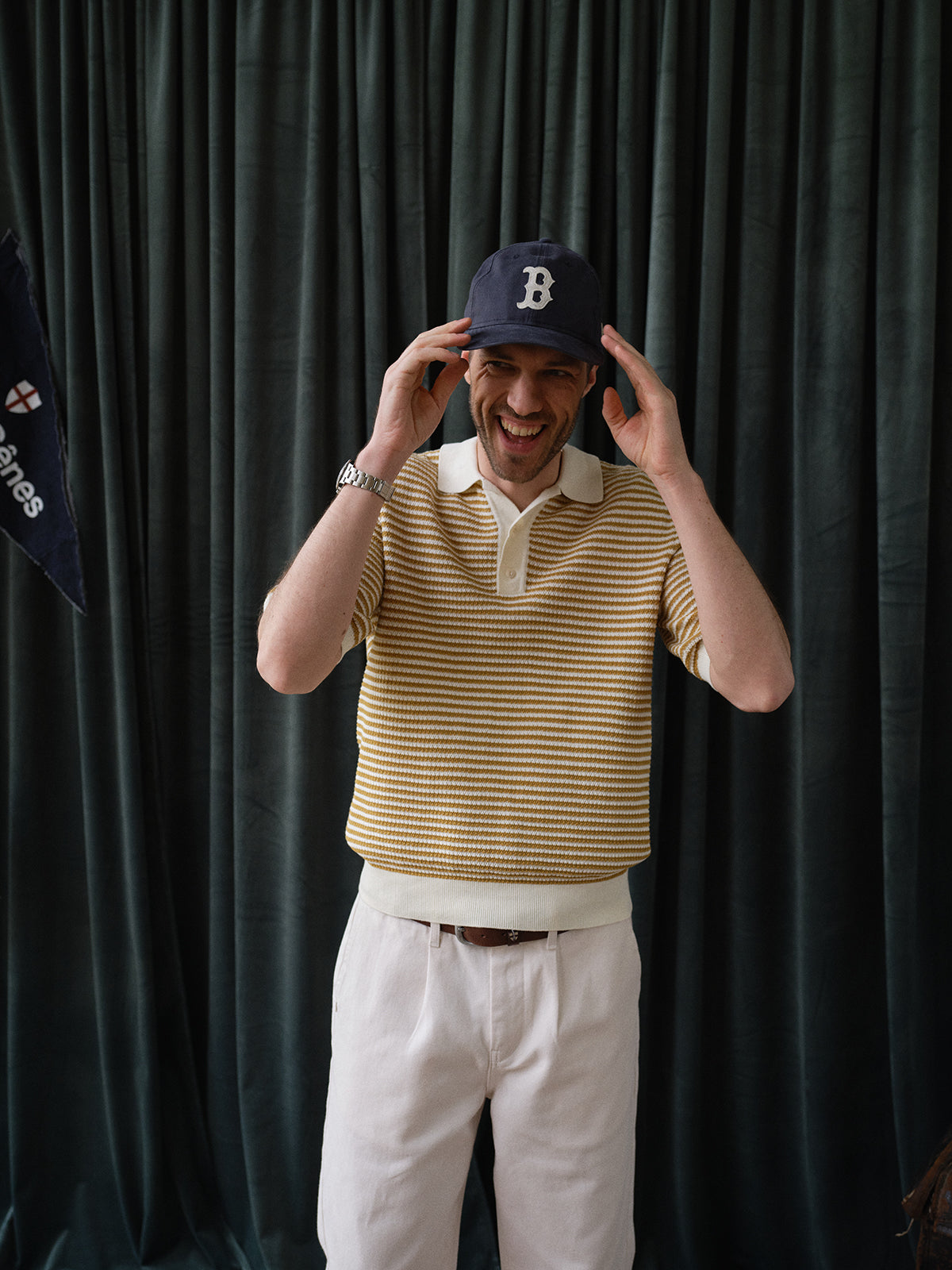 A man smiles while adjusting a navy cap, wearing the BGbob Knit Polo - Sunflower by Blue de Gênes—a yellow and white striped soft cotton knit polo—paired with white pants, standing before dark curtains.