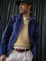 A man wearing the Blue de Gênes BGcanopy Jacket in Navy Peony, a striped yellow and white sweater, beige pants, and a navy cap stands before dark green curtains, looking left with one hand in his pocket.