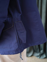 Close-up of the lower half of the BGcanopy Jacket – Navy Peony by Blue de Gênes, featuring a pocket and drawstring detail, worn over light-colored pants. A blurred dark green curtain is in the background.