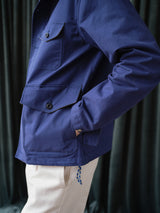 A person wears the BGcanopy Jacket in Navy Peony by Blue de Gênes, featuring front pockets, paired with beige pants, standing with one hand in their pocket before dark curtains.
