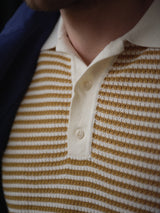 Close-up of a person wearing the Blue de Gênes BGbob Knit Polo - Sunflower, featuring cream and mustard stripes, a collared neckline, short sleeves, and a cream button placket. The lower face and part of a dark jacket are also visible.