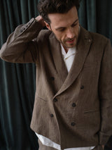 A man wears the Blue de Gênes BGcricket Lin Blazer in Grey Mel over a white shirt, standing with one hand behind his head and looking down, in front of dark pleated curtains.