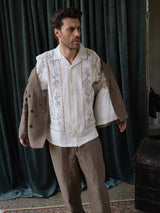 A man stands indoors in Blue de Gênes BGpalco Lin Pants in Grey Mel, paired with an embroidered white shirt and a jacket over his shoulders. Dark curtains and part of a trunk appear in the background.