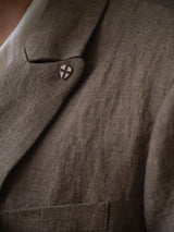 Close-up of the BGcricket Lin Blazer - Grey Mel by Blue de Gênes, a brown relaxed fit blazer with a small enamel pin featuring a black cross on a white shield. A white shirt is partially visible underneath.