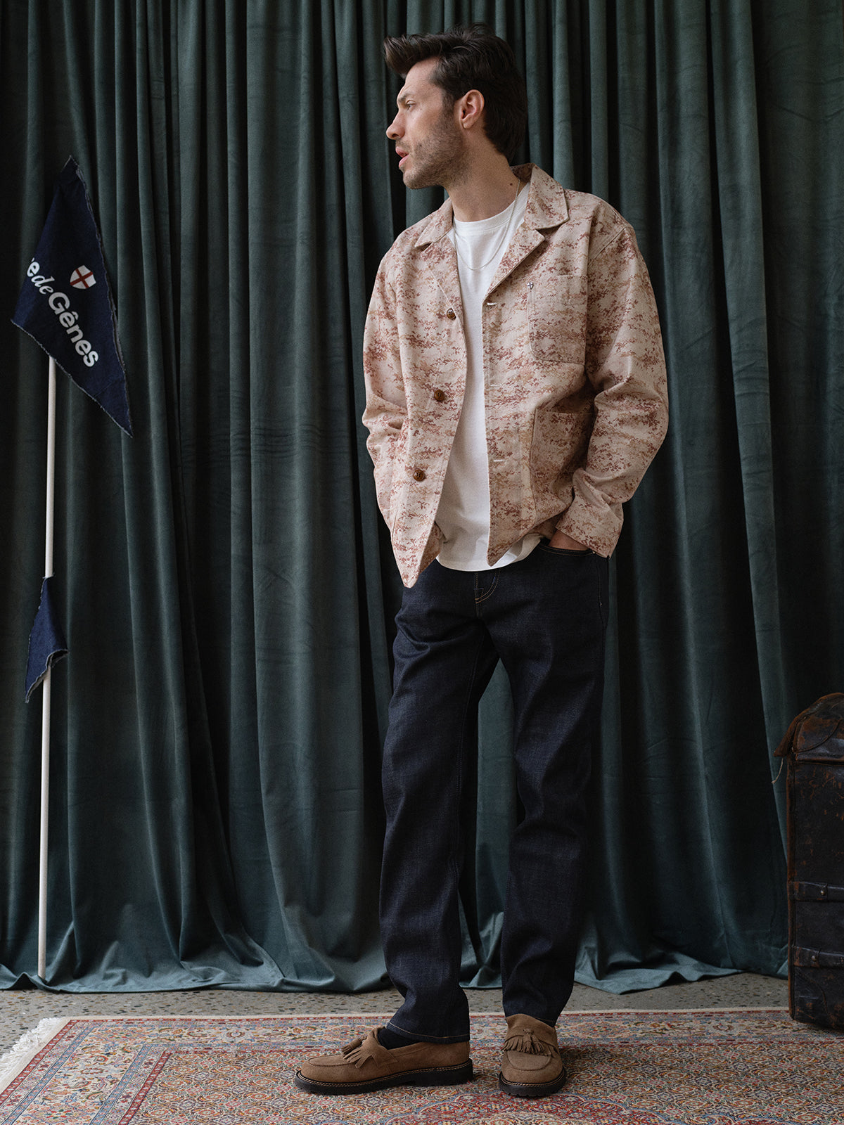 A man stands on a patterned rug before dark green curtains, wearing a white t-shirt, beige patterned jacket, BGgiacomo Selvedge V2 Jeans - Raw Denim by Blue de Gênes, and brown shoes. A flag with berlGens text appears on the left.