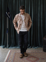 A man stands indoors on a patterned rug, wearing the Blue de Gênes BGkendrick Print Jacket - Multi Pattern over a white t-shirt, with dark jeans and brown shoes. Dark green curtains and a flag with velGenes text are visible in the background.
