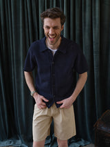 A smiling man wears Blue de Gênes BGcopple Rodas Shorts in Pale Khaki with a navy blue knitted short-sleeve shirt, standing before dark green curtains with his hands in his pockets.