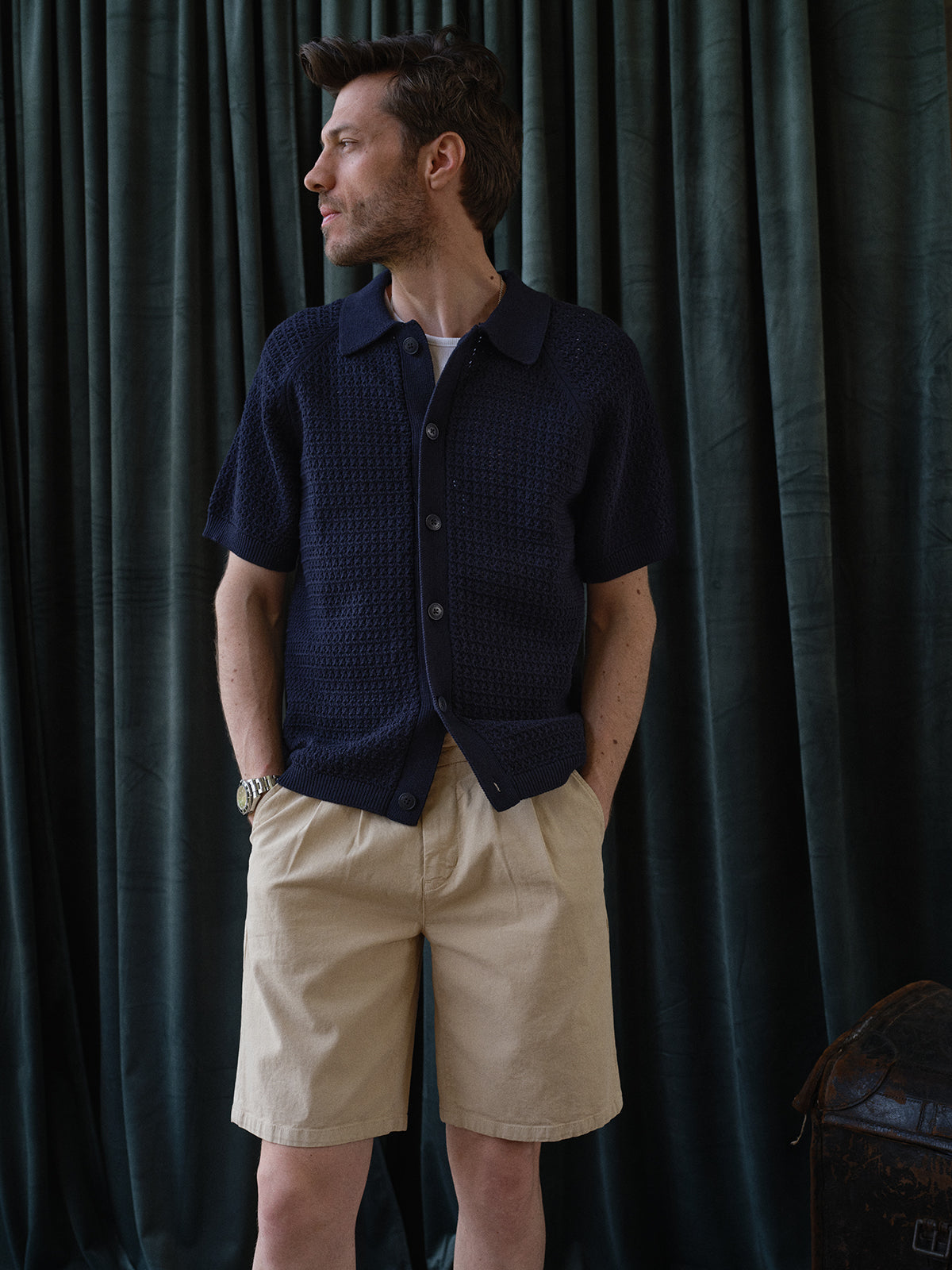 A man stands before dark green curtains, hands in pockets, wearing the Blue de Gênes BGbilly S/S Cardigan in Dark Navy over a dark tee, beige shorts, and a watch, gazing to the side.