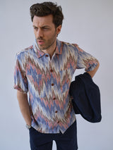 A man with wavy dark hair wears the Blue de Gênes BGponti Kala S/S Shirt—multi-patterned in blue, red, and beige. He holds a navy jacket over his left arm and gazes seriously to the side against a plain backdrop.