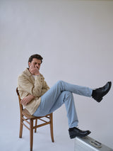 A man sits on a wooden chair with one leg crossed, wearing a beige jacket, black shoes, and Blue de Gênes BGpaulo Pavia Pant in Blue Fog. He rests his hand on his face; a silver suitcase sits before him against a plain background.