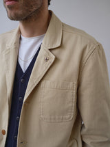 A man wears the Blue de Gênes BGnorly Rodas Blazer in pale khaki over a navy cardigan and white T-shirt, accessorized with a small lapel pin and thin chain necklace; image is cropped to the upper torso.