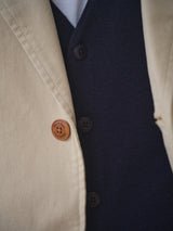 Close-up of a person wearing the BGnorly Rodas Blazer in Pale Khaki by Blue de Gênes, crafted from a cotton-hemp blend with brown buttons, layered over a dark navy vest and white shirt. The textures and details are clearly visible.