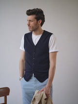 A man with short brown hair wears a white t-shirt, light blue pants, and the Blue de Gênes Zorba Knit Waistcoat in navy as he stands against a plain background, holding a beige jacket and looking to the side.