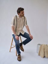 A man in a short-sleeved, striped shirt, blue jeans, and brown shoes sits on a stool. Next to his beige jacket on the floor are a BGwalter S/S Cardigan - Chalk by Blue de Gênes and a silver suitcase.
