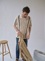 A man wears the Blue de Gênes BGwalter S/S Cardigan in Chalk with black stripes and jeans indoors, holding a beige jacket. Nearby are a wooden stool and silver suitcase against a plain, light background.