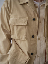 Close-up of a person wearing the BGhunter Vecchio Jacket in Pale Khaki by Blue de Gênes, featuring large flap pockets and detailed buttons, layered over a textured cream shirt and white T-shirt. Focus is on the jacket’s craftsmanship.