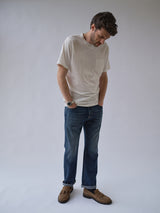 A man stands with his hands in his pockets, looking down, wearing a white T-shirt, Blue de Gênes BGmarco Rowan Jeans - Used with a relaxed fit and classic 5-pocket design, and brown loafers against a plain light background.
