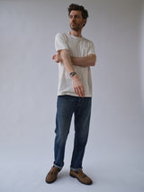 A man with short hair stands against a plain background, wearing a white t-shirt, Blue de Gênes BGmarco Rowan Jeans - Used (relaxed fit), brown shoes, and a watch, with one arm crossed over the other.