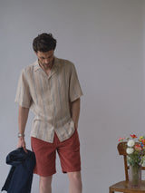 A man stands indoors wearing the Blue de Gênes BGcody Fling Shirt in Pale Khaki with red shorts, holding a dark jacket. A wooden chair with a vase of colorful flowers sits beside him against a plain background.