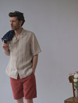 Wearing the Blue de Gênes BGcody Fling Shirt in pale khaki and red shorts, a man stands with a jacket over his shoulder, gazing to the side. Beside him, a wooden chair with flowers sits against a plain background.