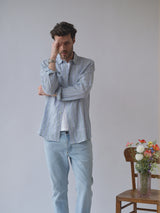 A man in the Blue de Gênes BGenrico Mida Shirt - Blue Stripe and light jeans stands thoughtfully with one arm crossed, hand to forehead. Beside him is a wooden chair holding a glass vase of colorful flowers against a plain backdrop.