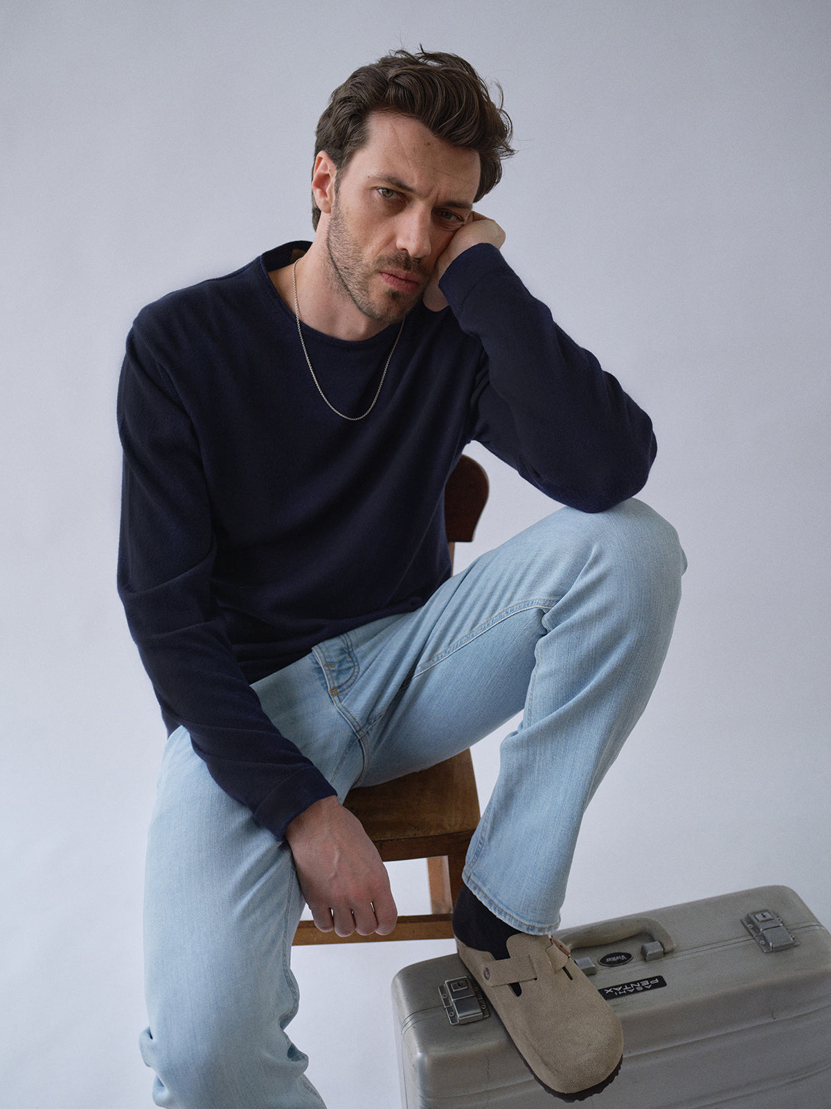 A man with short brown hair sits thoughtfully on a chair, resting his head on his hand. He wears the Blue de Gênes BGmonty Knit - Dark Navy, light blue jeans, beige slip-on shoes, a silver chain, and sits beside a gray suitcase.
