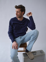 A man in a navy blue sweater and BGvinci Trueblue Jeans - Mid Blue Denim from Blue de Gênes sits with one leg raised, holding a card and looking to the side. A light gray suitcase is on the floor near his foot.
