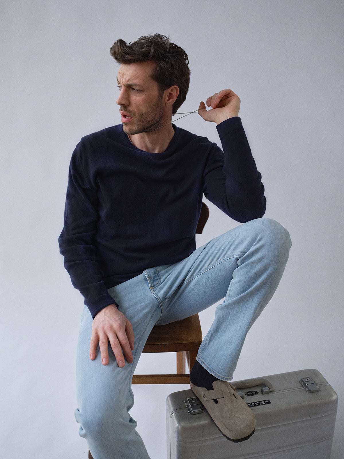 Wearing the BGmonty Knit in Dark Navy by Blue de Gênes and light blue jeans, a man sits on a chair with one leg over a silver suitcase, glancing left in surprise as he lifts a piece of paper in his hand.