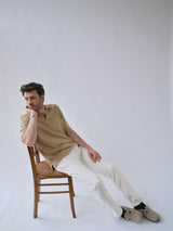 A man, dressed in the Blue de Gênes BGjohn Slubby Polo in Pale Khaki and white pants, sits sideways on a wooden chair, leaning back thoughtfully with his hand on his chin against a plain light background.
