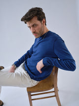 A man with wavy brown hair and a short beard sits sideways on a wooden chair, wearing the Blue de Gênes BGmonty Knit in Navy Peony and white pants, looking seriously at the camera against a plain light background.